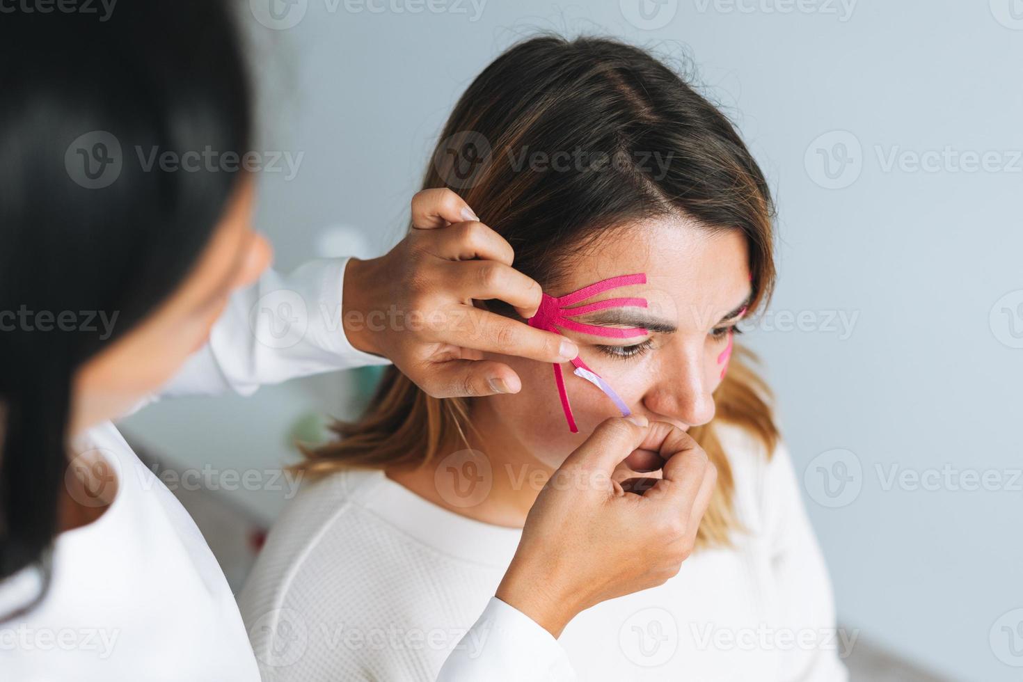 joven y hermosa mujer morena con el cosmetólogo médico de pelo largo hace cintas faciales para el paciente en el consultorio. las mujeres jóvenes realizan procedimientos antienvejecimiento con ayuda de cinta kinesio foto