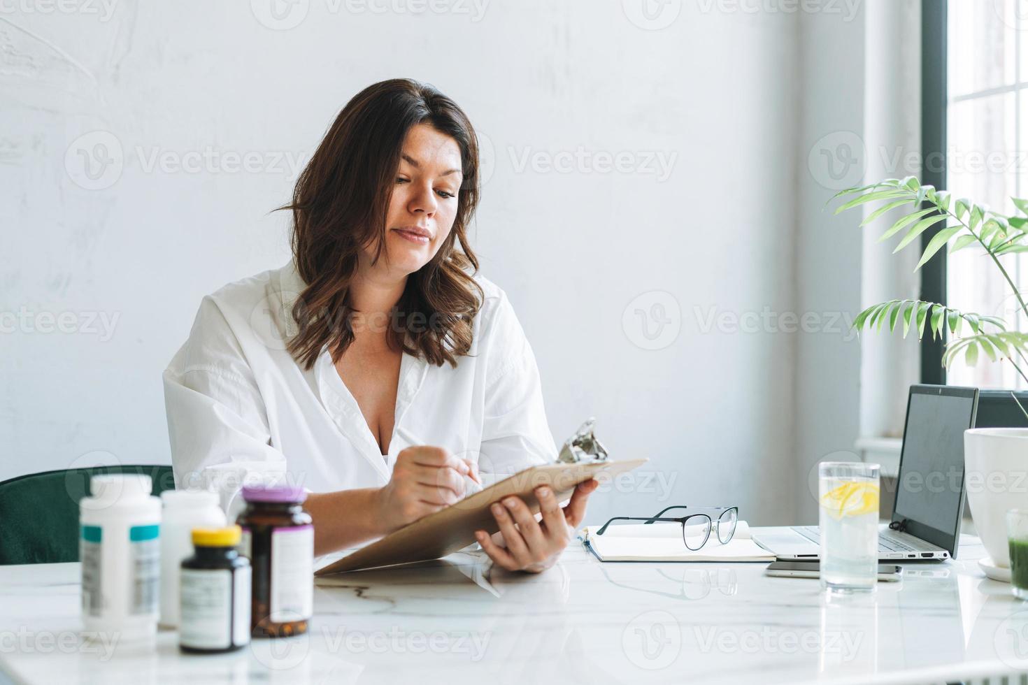 joven morena sonriente doctora nutricionista de talla grande con camisa blanca trabajando en una laptop en una oficina moderna y luminosa. el médico prescribe una receta de medicamentos y vitaminas en la clínica foto
