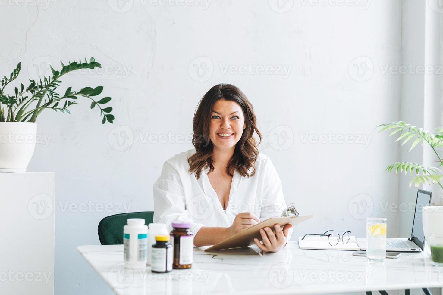 joven morena sonriente doctora nutricionista de talla grande con camisa blanca trabajando en una laptop en una oficina moderna y luminosa. el médico prescribe una receta de medicamentos y vitaminas en la clínica foto