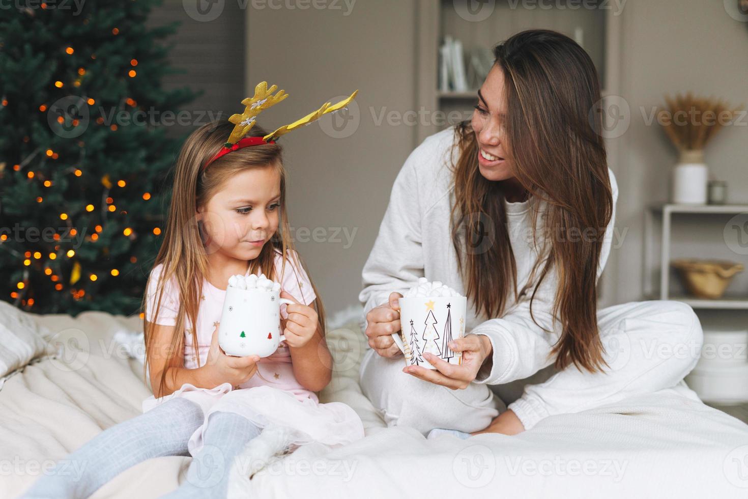 Cute girl with mother on bed in room with Christmas tree. Portrait of child in the rim with deer horns in cottage house in christmas time, happy New Year. Happy family with cocoa and marshmallow photo