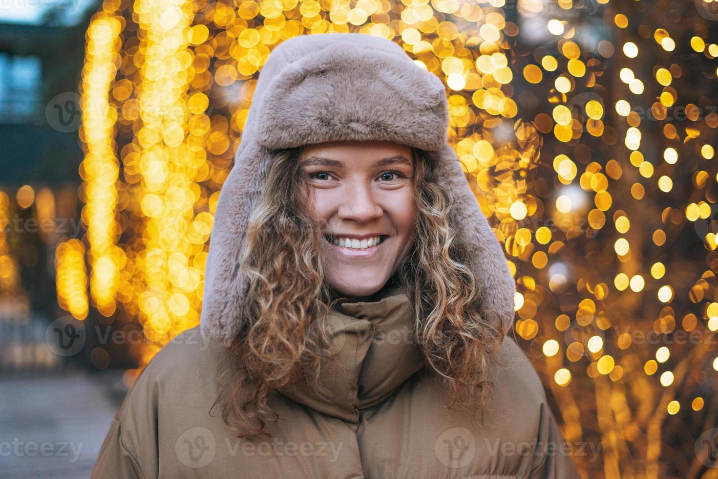 Portrait of young smiling woman with curly hair in fur hat in the winter street decorated with lights photo