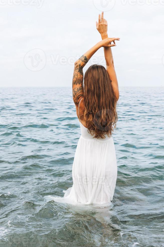 joven mujer despreocupada con el pelo largo vestido de blanco disfrutando de la vida en la playa, gente por detrás foto