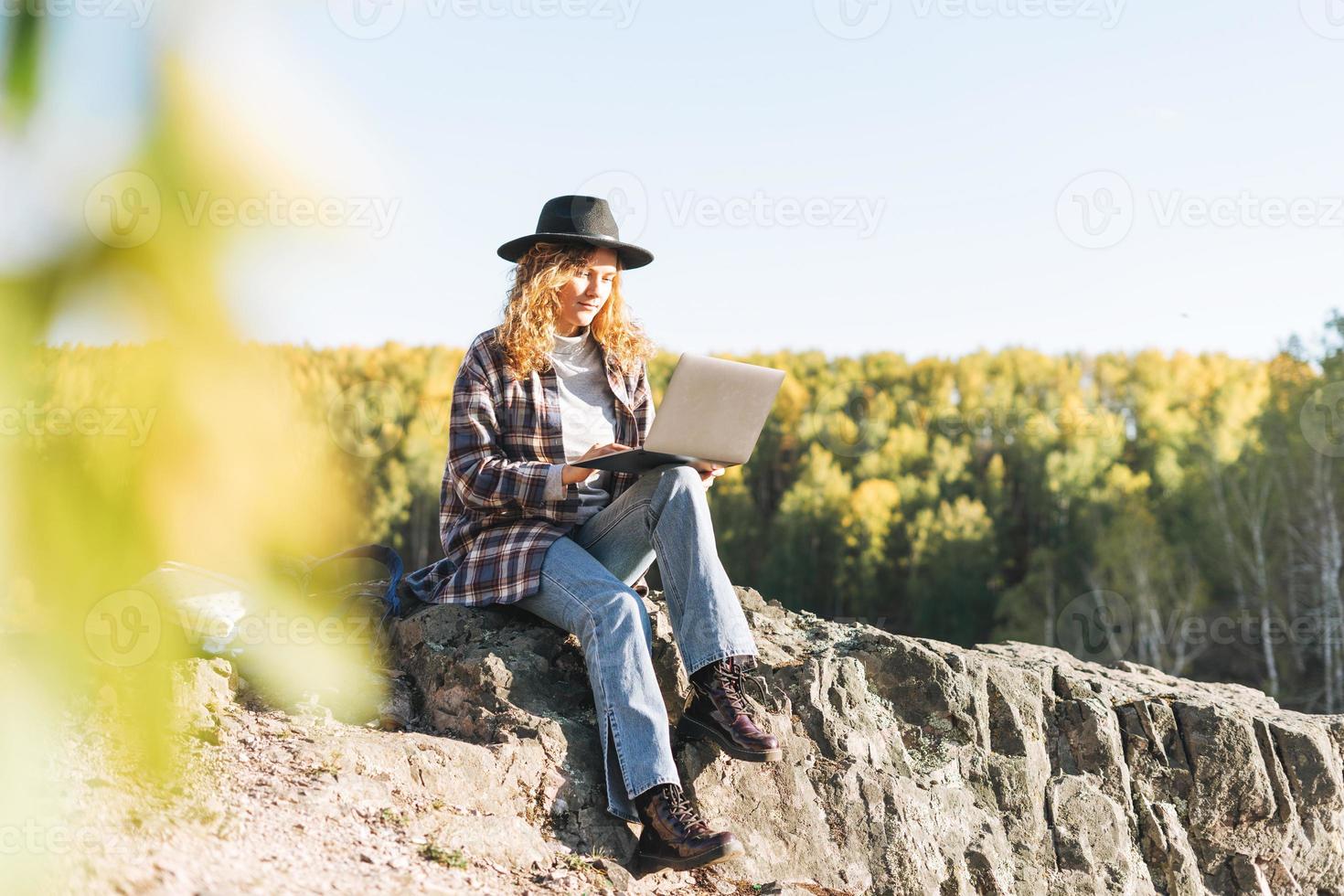 joven mujer hermosa con cabello rizado en sombrero de fieltro y camisa a cuadros usando una computadora portátil en el fondo de las montañas, caminando en la naturaleza otoñal, concepto de freelance foto