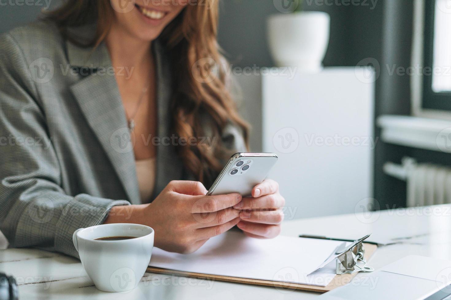 Crop photo of Young smiling brunette woman with long hair in stylish suit using mobile phone in hands working in the bright modern office, woman send message by smartphone