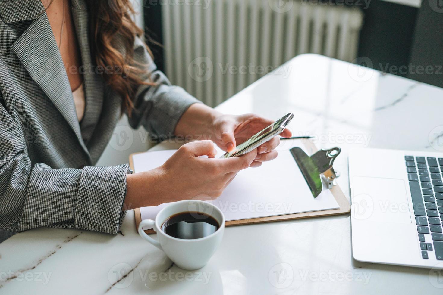 Crop photo of Young smiling brunette woman with long hair in stylish suit using mobile phone in hands working in bright modern office, woman send message by smartphone