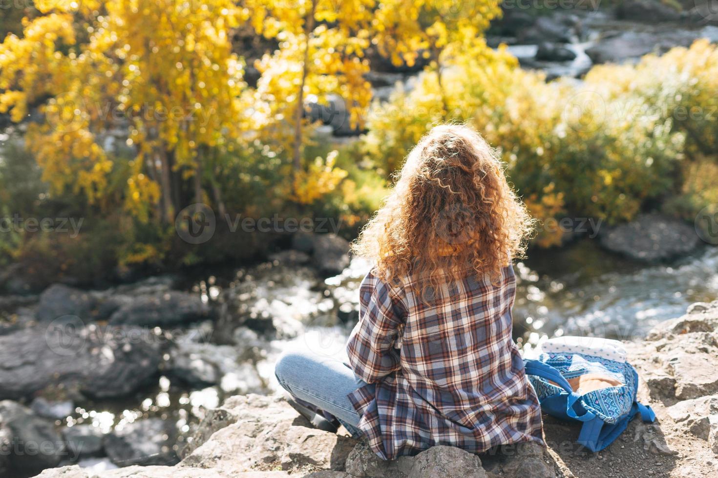 Young beautiful woman with curly hair in plaid shirt, jeans looks at magic view of mountains and river, hiking on the autumn nature, people from behind photo