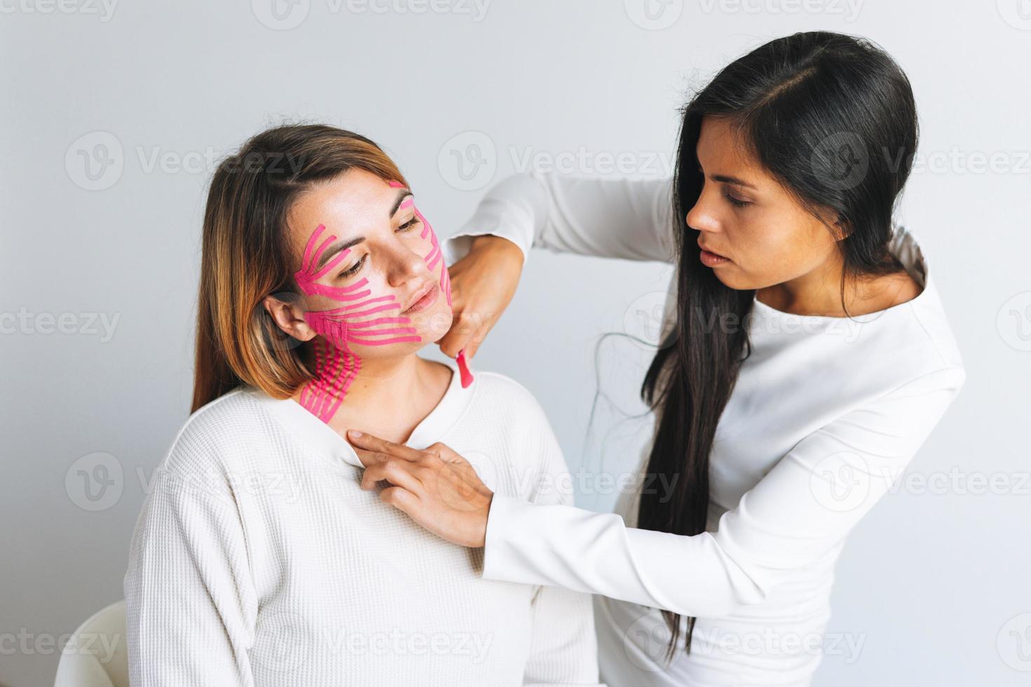 joven y hermosa mujer morena con el cosmetólogo médico de pelo largo hace cintas faciales para el paciente en el consultorio. las mujeres jóvenes realizan procedimientos antienvejecimiento con ayuda de cinta kinesio foto