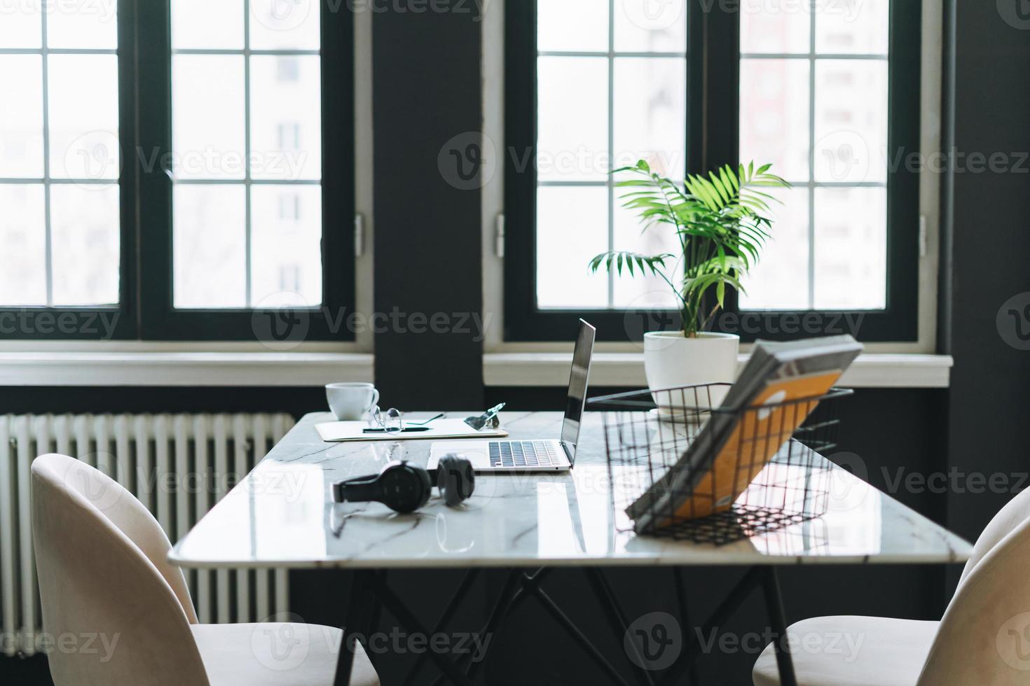 lugar de trabajo con taza de café, notas, computadora portátil abierta, anteojos y teléfono inteligente en una mesa de mármol blanco en la oscura y moderna sala de oficinas, interior de negocios foto