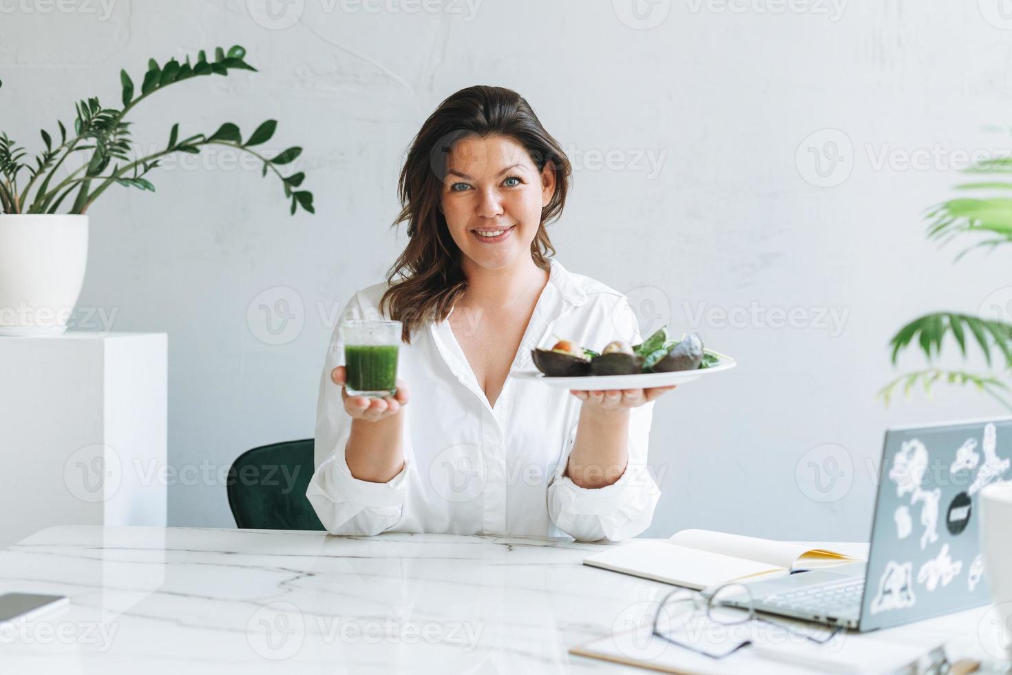 joven morena sonriente doctora nutricionista de talla grande con camisa blanca con comida verde saludable en la moderna y luminosa sala de oficina. el médico se comunica con el paciente en línea foto