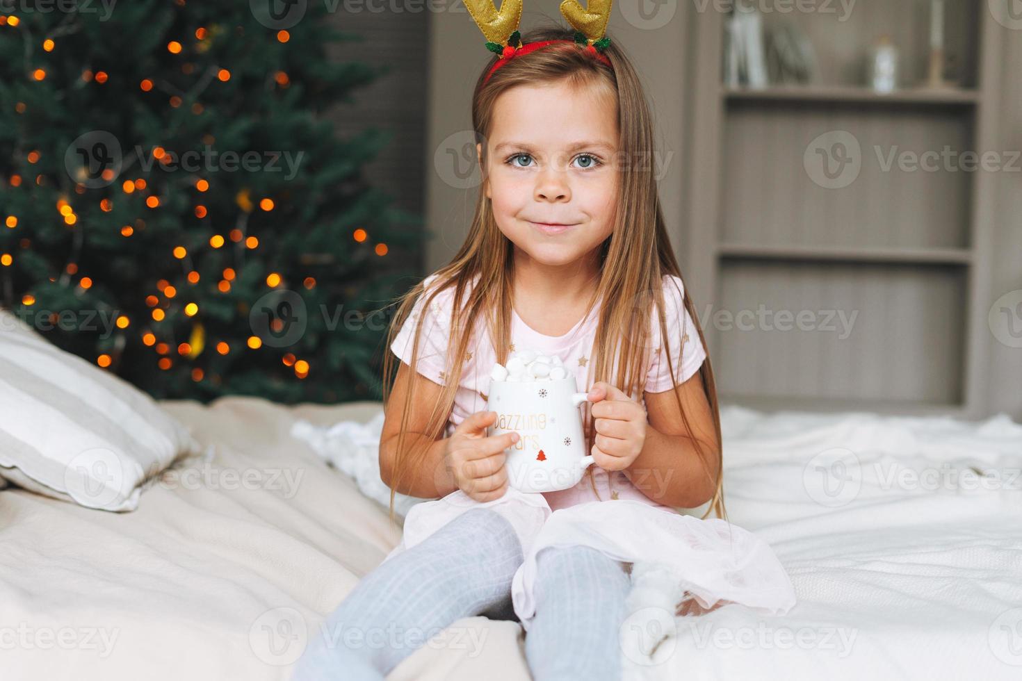 Linda niña graciosa y sonriente con vestido rosa bebiendo cacao sentada en la cama en la habitación con árbol de Navidad. retrato de niño en el borde con cuernos de ciervo en casa de campo en Navidad, feliz año nuevo foto