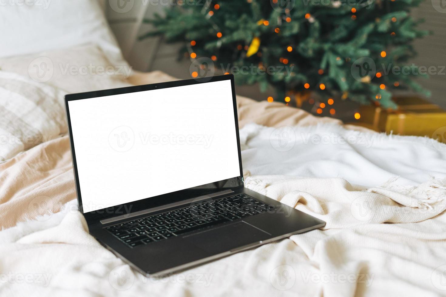 Opened laptop with white screen on bed in bedroom with decorated Christmas tree at home, mockup photo