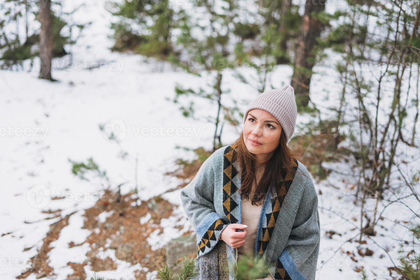 Portrait of young brunette beautiful woman in hat and grey poncho in the winter forest photo