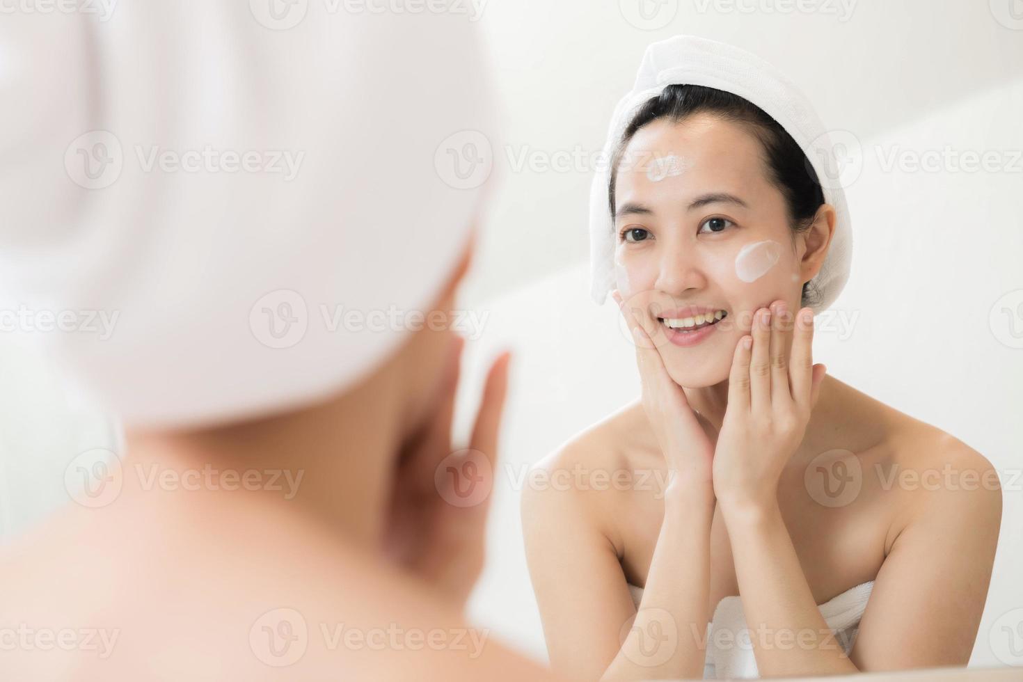 feliz joven asiática aplicando lociones faciales mientras usa una toalla y toca su cara en el baño foto