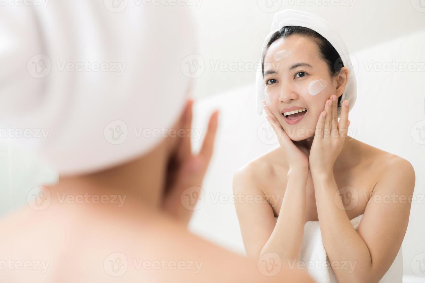 Happy young Asian woman applying face lotions while wearing a towel and touching her face in bathroom photo