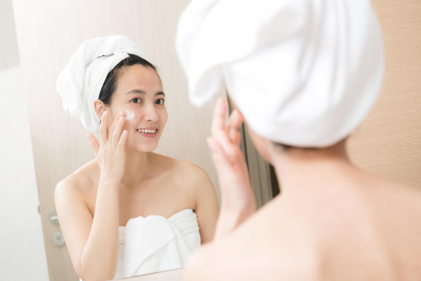 Happy young Asian woman applying face lotions while wearing a towel and touching her face in bathroom photo