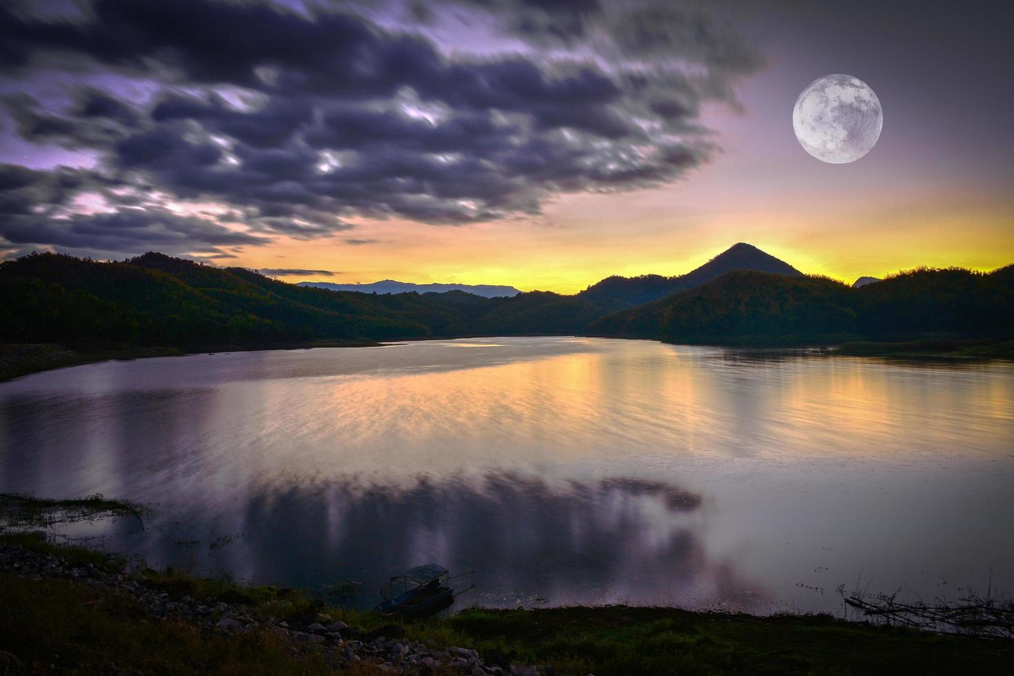 paisaje lago río naturaleza con luna llena puesta de sol cielo amarillo y morado nubes oscuras moviéndose en silueta montaña foto