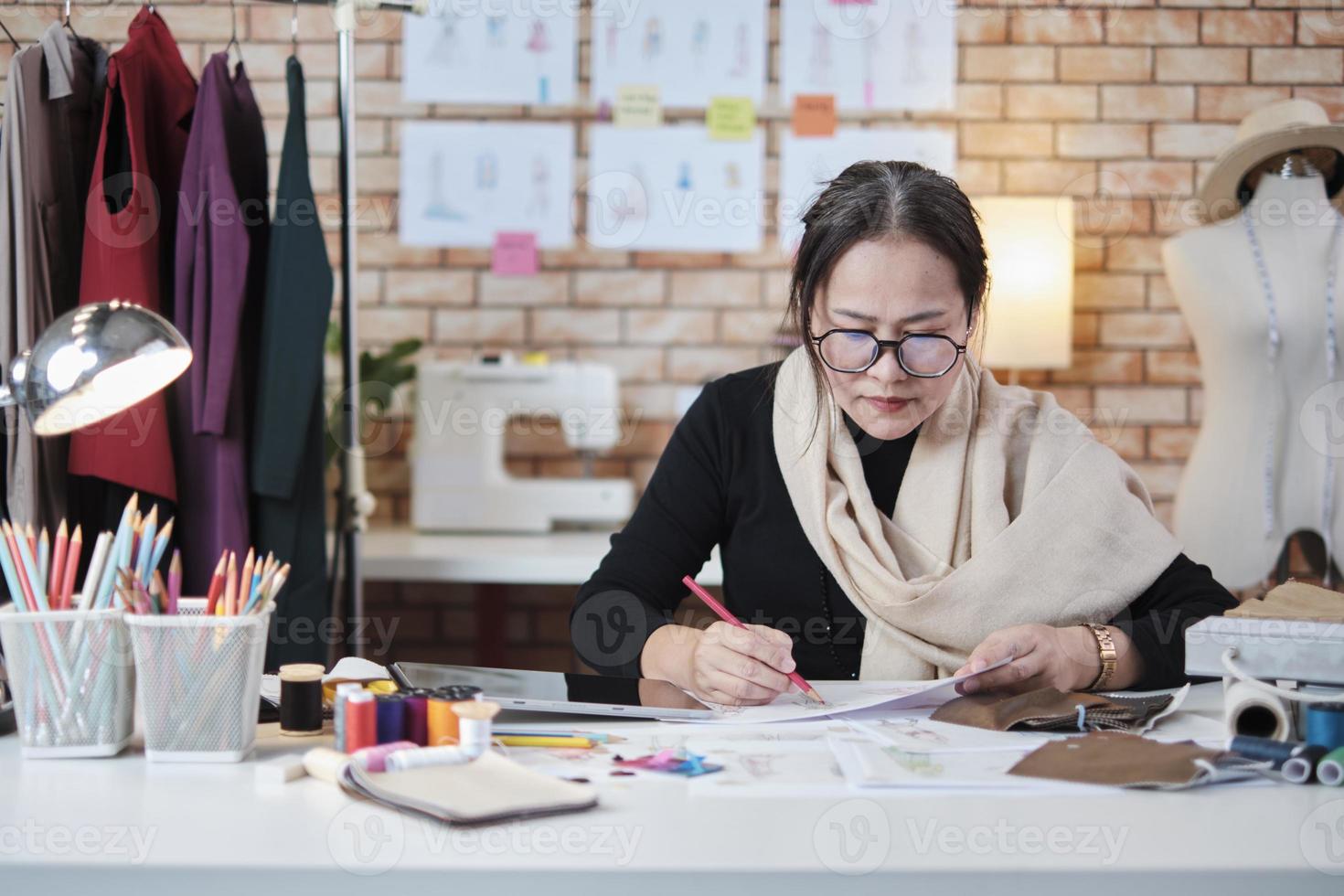 Asian middle-aged female fashion designer working in a studio by idea drawing sketches with colorful thread and sewing for a dress design collection, professional boutique tailor SME entrepreneur. photo