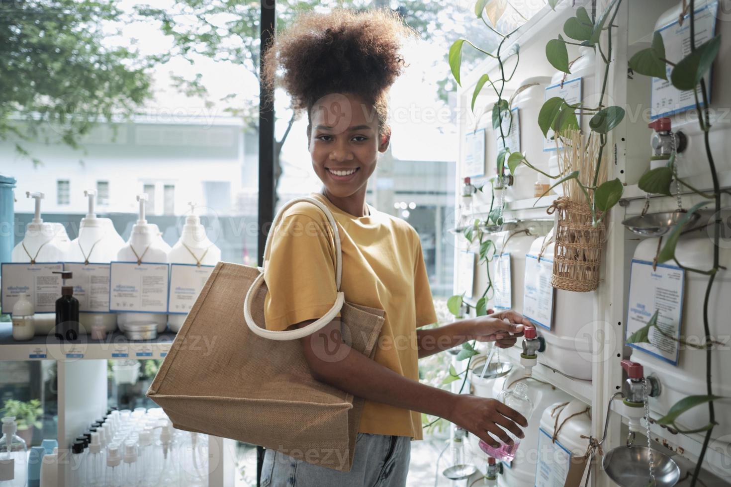 A Black female customer fills liquid soap into recycling jar from reusable containers in a zero-waste and refill store, smiles, and looks at camera, happy shopping in an environment-friendly shop. photo