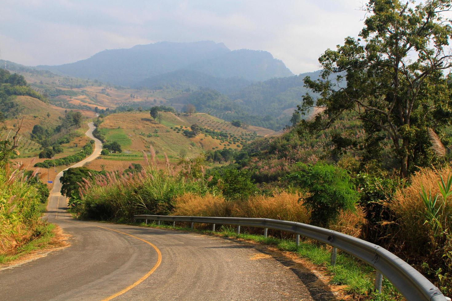 carretera asfaltada curva que baja y sube a la colina junto con montañas y nubes fondo de cielo azul en el parque forestal phu langka, phayao, tailandia. belleza de lo natural. hermoso camino, viaje y calle. foto