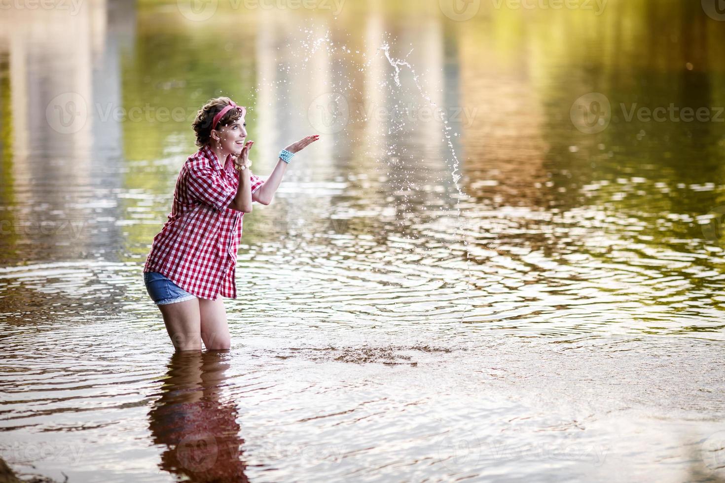 hermosa joven en una camisa a cuadros y pantalones cortos de mezclilla en estilo pin-up permanece en el agua del río foto