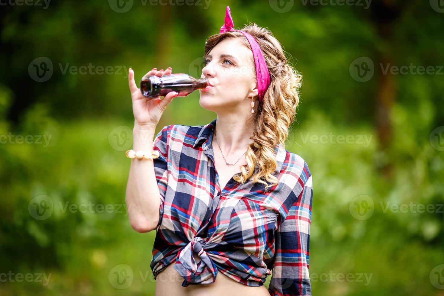 beautiful young girl in a plaid shirt and short denim shorts in pin-up style in forest drinks photo