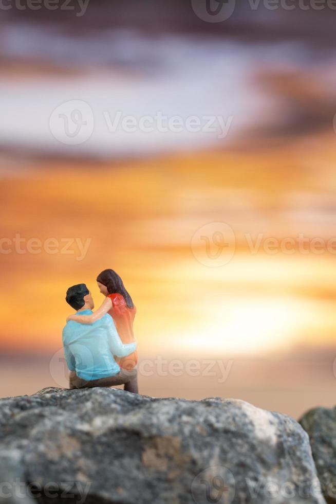 gente en miniatura, pareja sentada en una playa de mar con fondo de puesta de sol foto