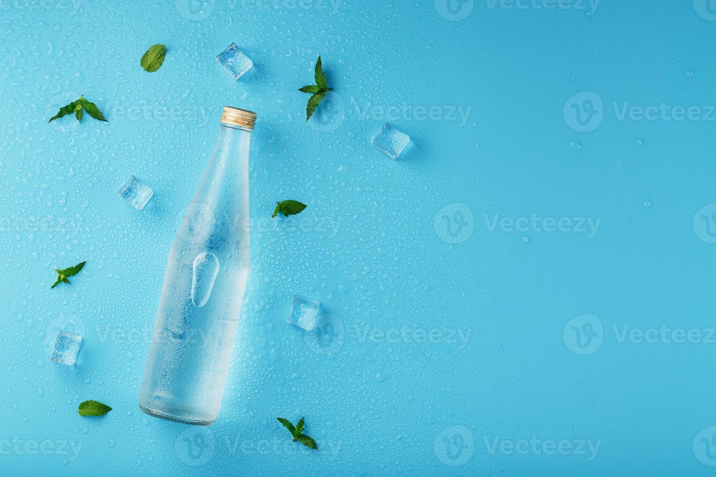 una botella de agua helada, cubitos de hielo, gotas y hojas de menta sobre un fondo azul. foto