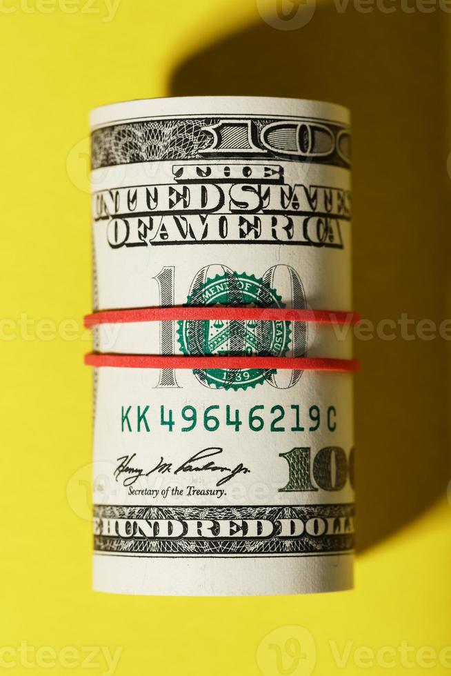 A roll of hundred-dollar American bills is tied with a red elastic band on a yellow background. photo