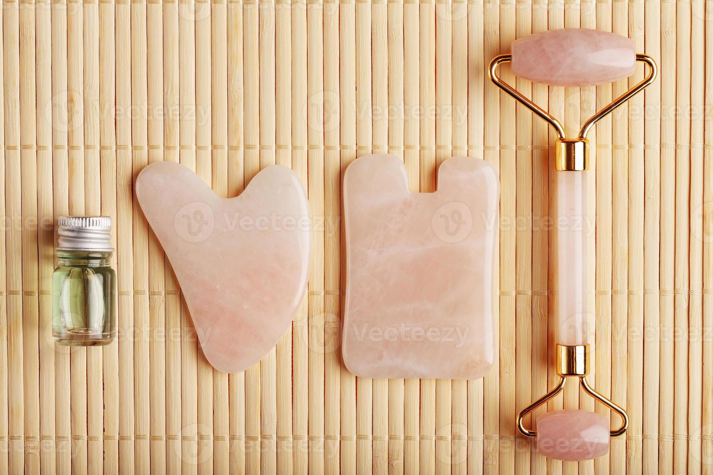 un conjunto de herramientas para la técnica de masaje facial gua sha fabricado en cuarzo rosa natural. rodillo, piedra de jade y aceite en un frasco de vidrio, sobre un fondo de paja para el cuidado de la cara y el cuerpo. foto