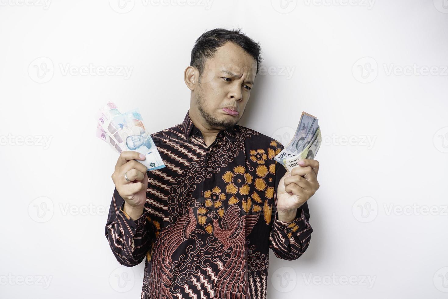 A confused young Asian man is wearing batik shirt and holding cash money in Indonesian rupiah isolated by white background photo