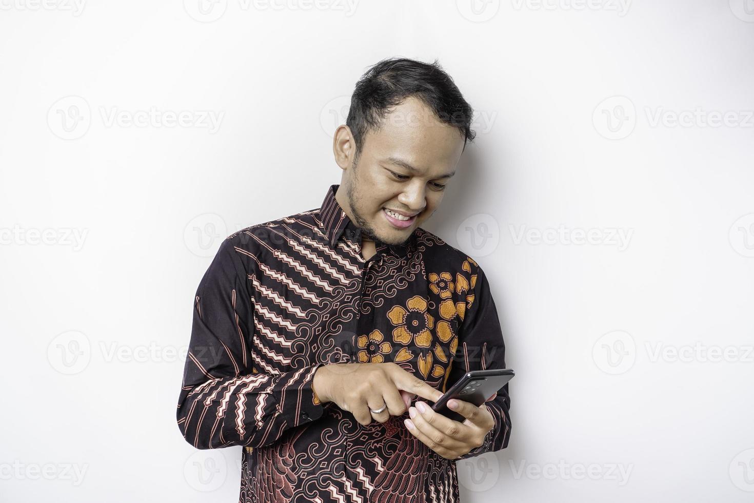 A portrait of a happy Asian man wearing batik shirt and holding his phone, isolated by white background photo