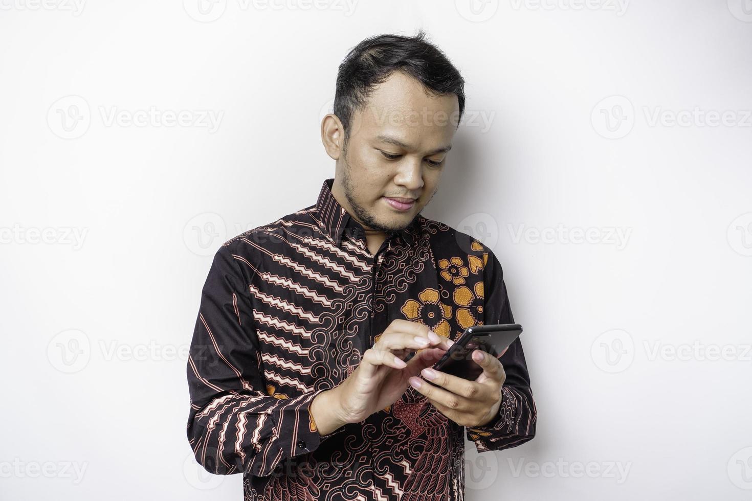 A portrait of a happy Asian man wearing batik shirt and holding his phone, isolated by white background photo