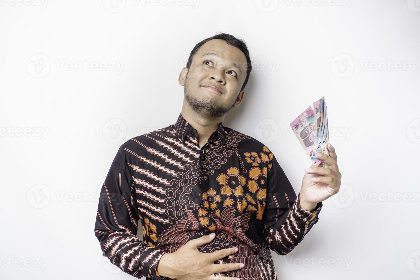 Photo of a hungry excited young Asian man holding money in Indonesian Rupiah and wondering what to order yummy food isolated on white color background