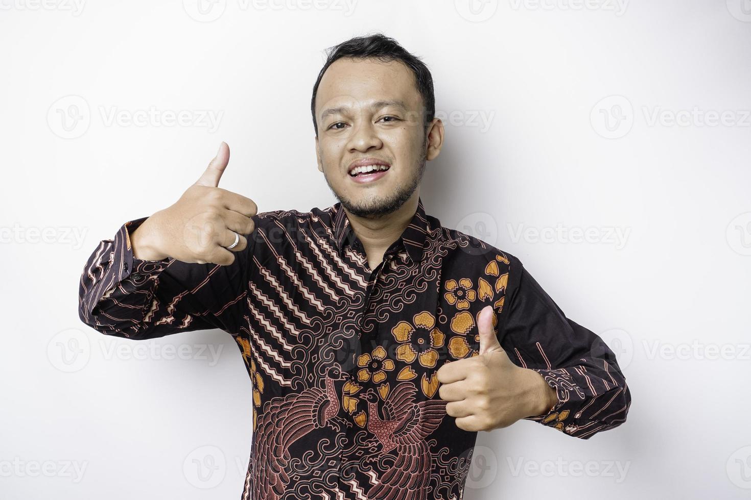 Excited Asian man wears batik shirt, gives thumbs up hand gesture of approval, isolated by white background photo