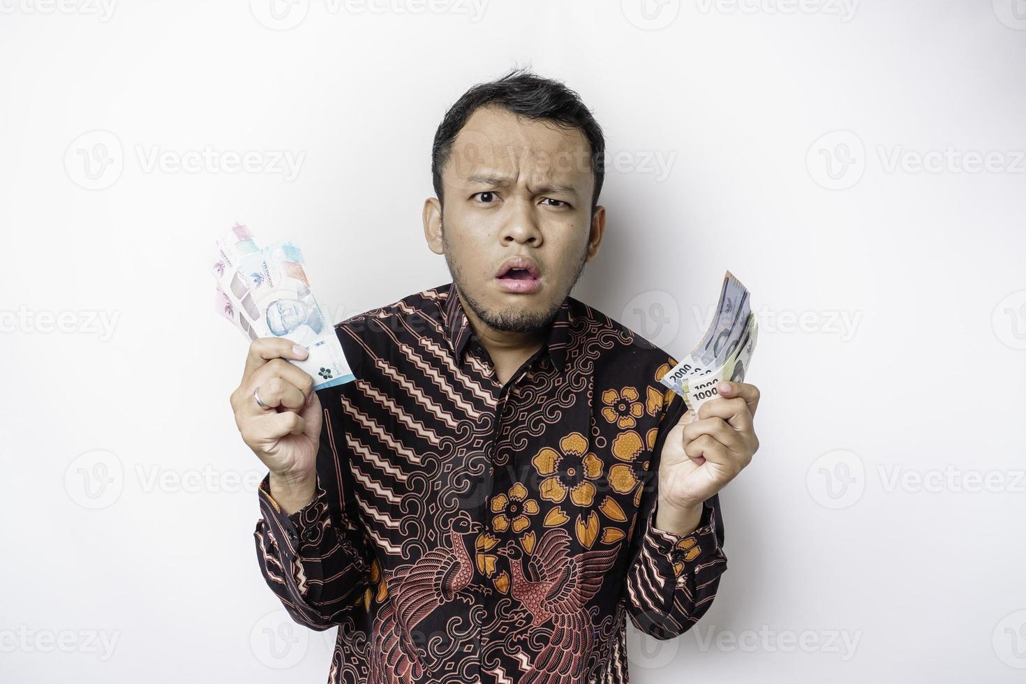 A confused young Asian man is wearing batik shirt and holding cash money in Indonesian rupiah isolated by white background photo