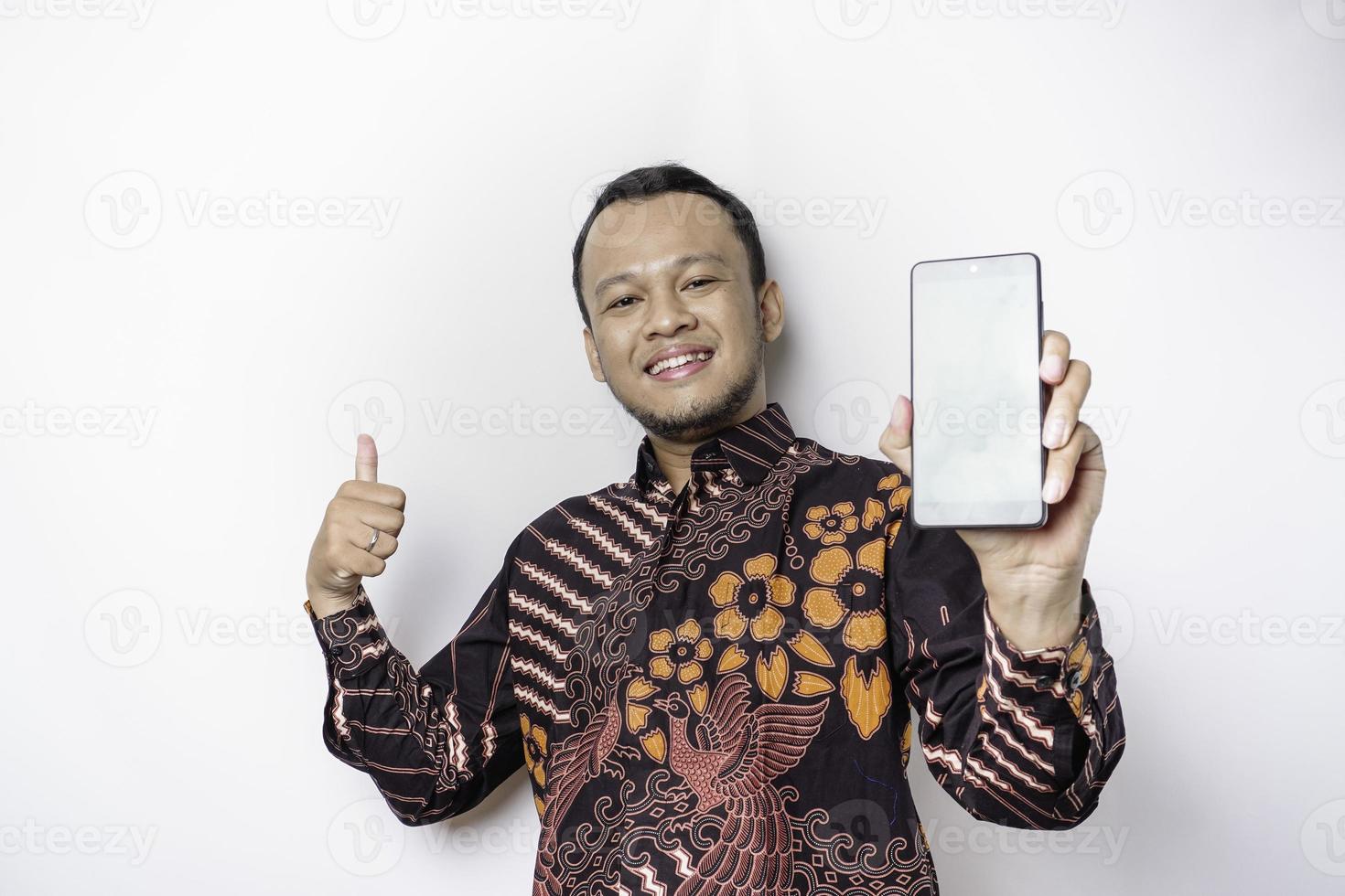 un retrato de un hombre asiático sonriente que lleva un pantalones de batik y muestra una pantalla verde en su teléfono, aislado de fondo blanco foto