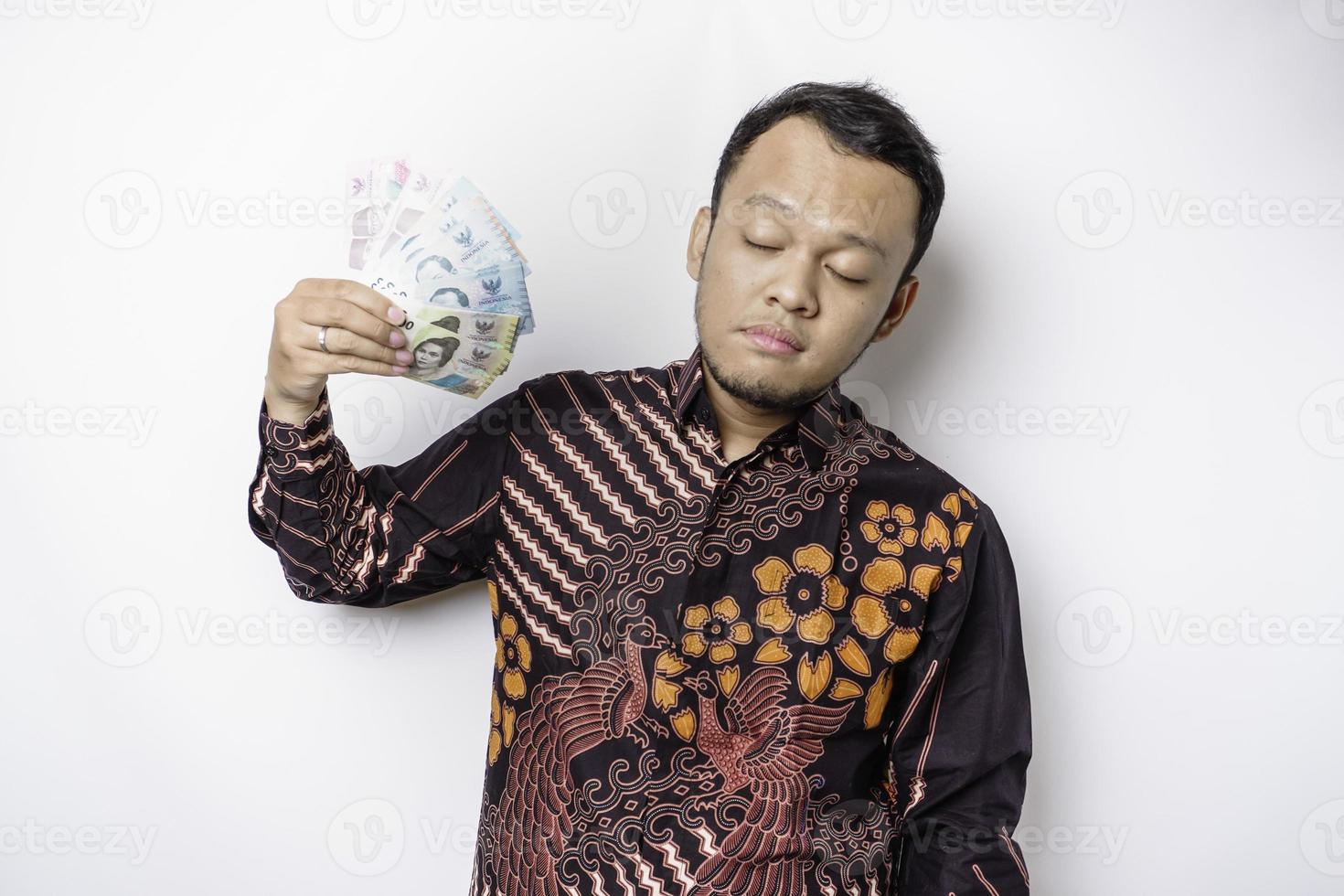 Young Asian man wears batik shirt holding fan of cash money in Indonesian rupiah banknotes doing isolated on white background studio portrait photo