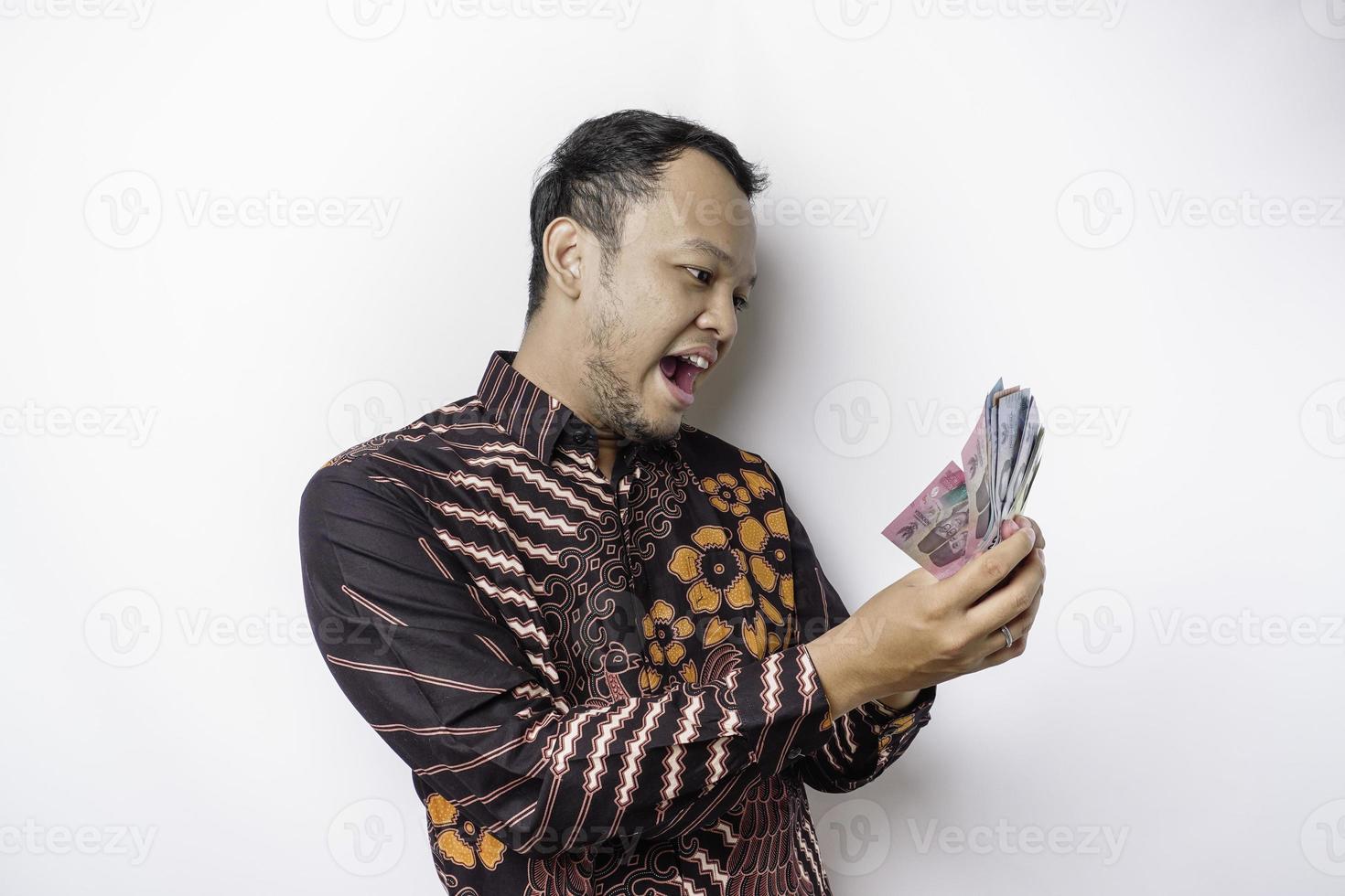 A happy young Asian man is wearing batik shirt and holding cash money in Indonesian rupiah isolated by white background photo