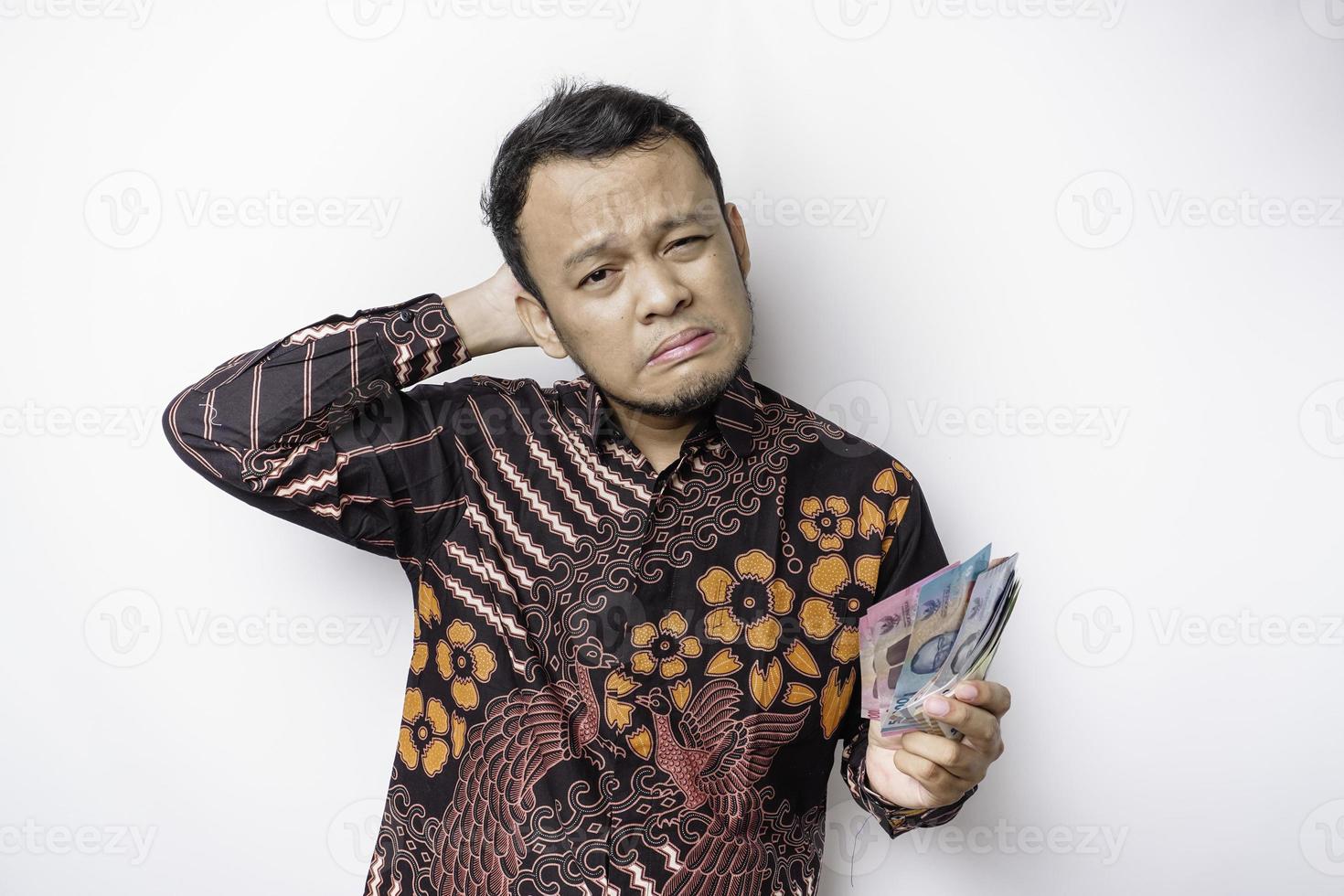 A stressed Asian man wearing batik shirt and holding money in Indonesian Rupiah isolated by white background looks depressed photo