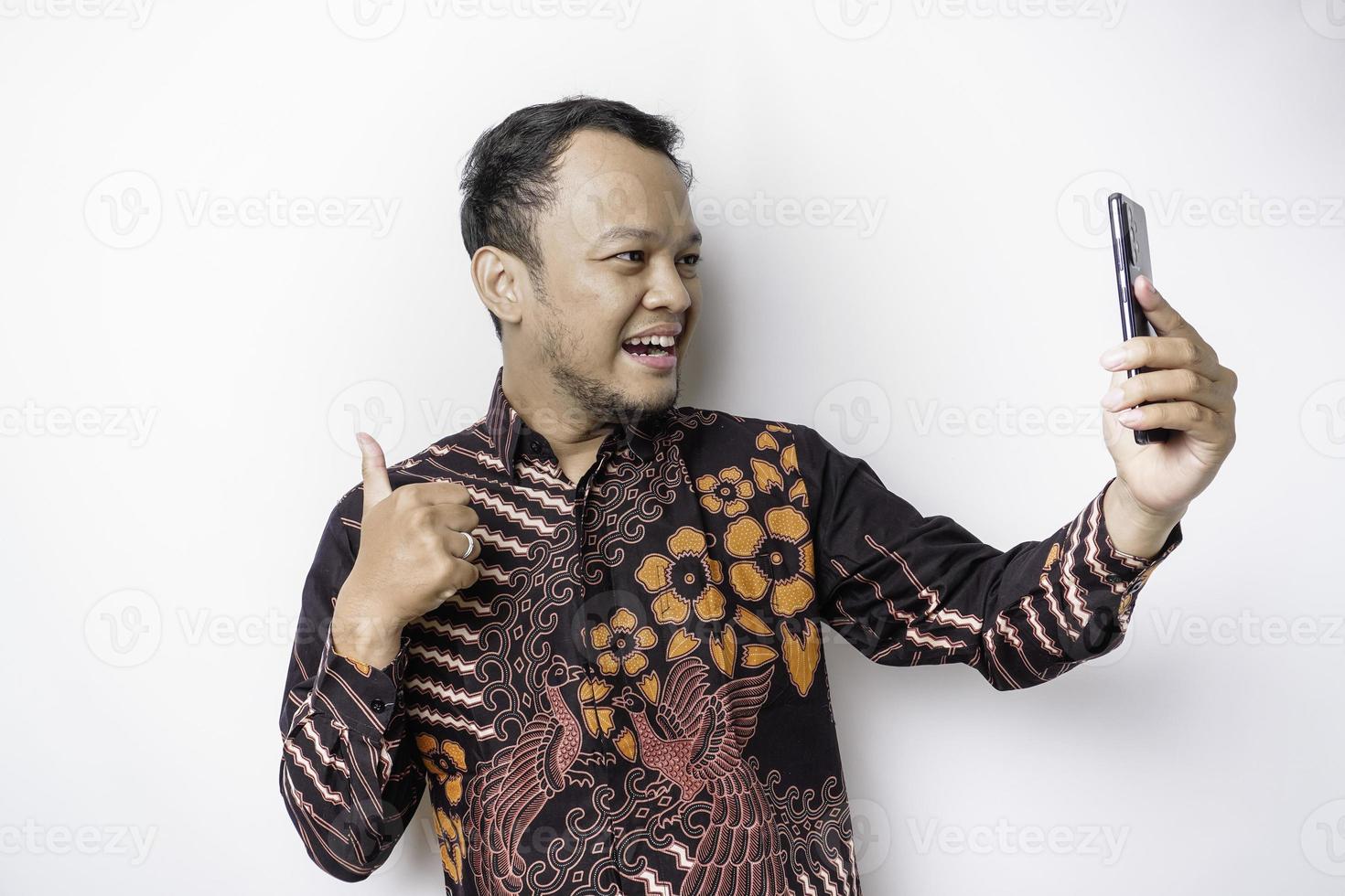 A portrait of a happy Asian man wearing batik shirt and holding his phone, isolated by white background photo