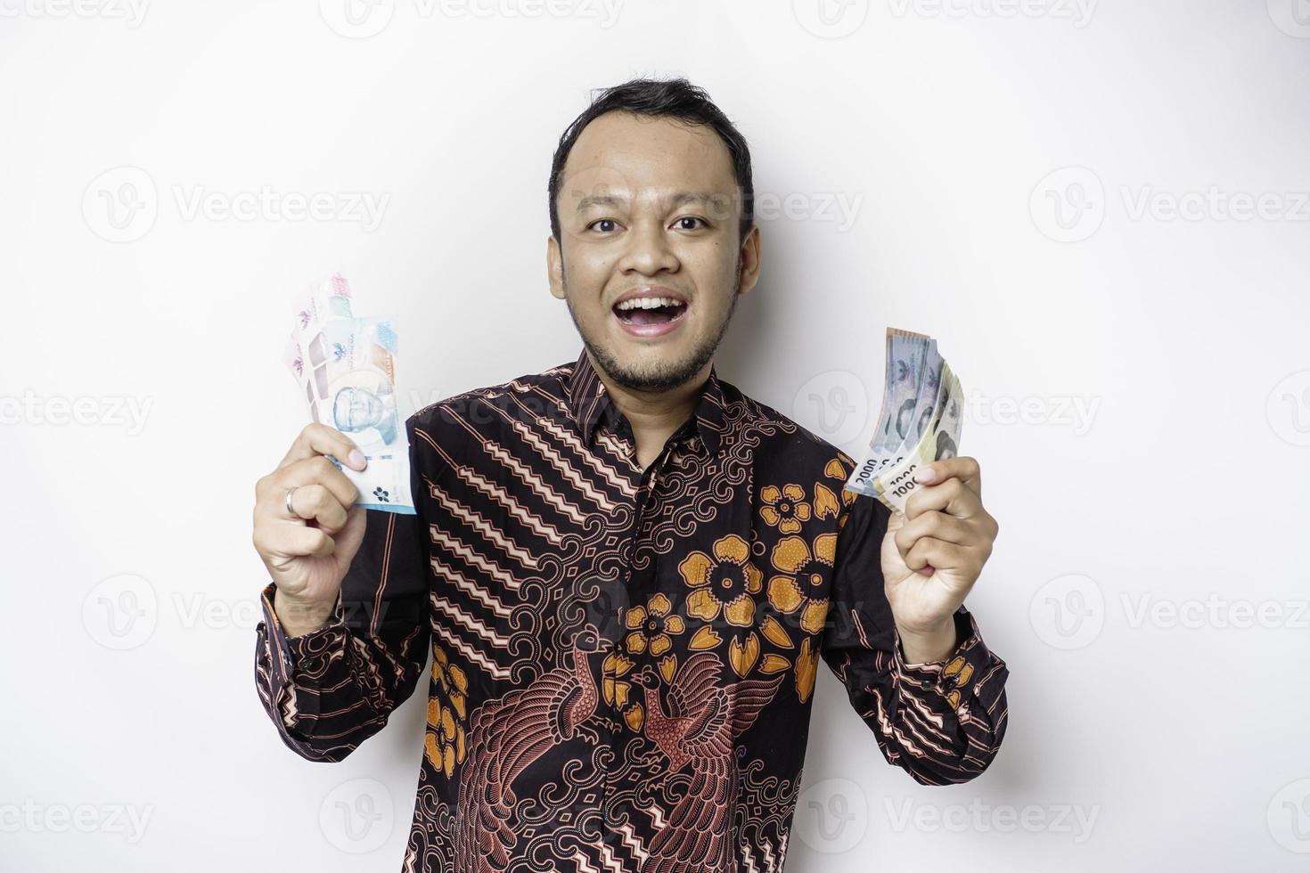 A happy young Asian man is wearing batik shirt and holding cash money in Indonesian rupiah isolated by white background photo