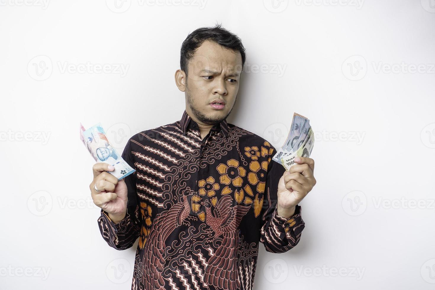 un joven asiático confundido lleva un pantalones batik y tiene dinero en efectivo en rupias indonesias aislado de fondo blanco foto