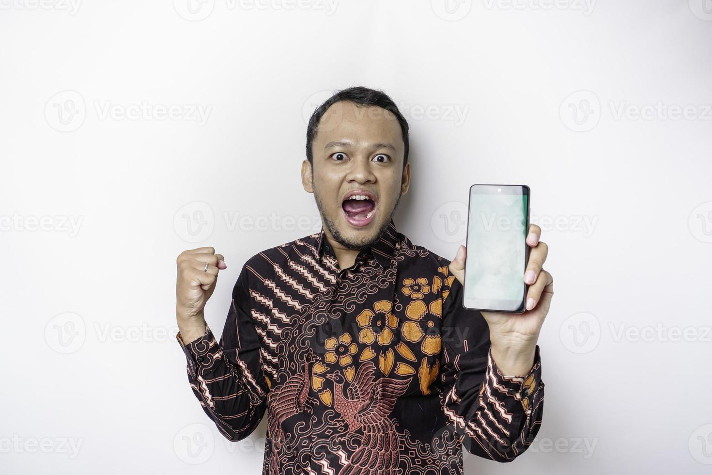 A happy young Asian man wearing batik shirt showing successful expression showing copy space on his phone isolated by white background photo