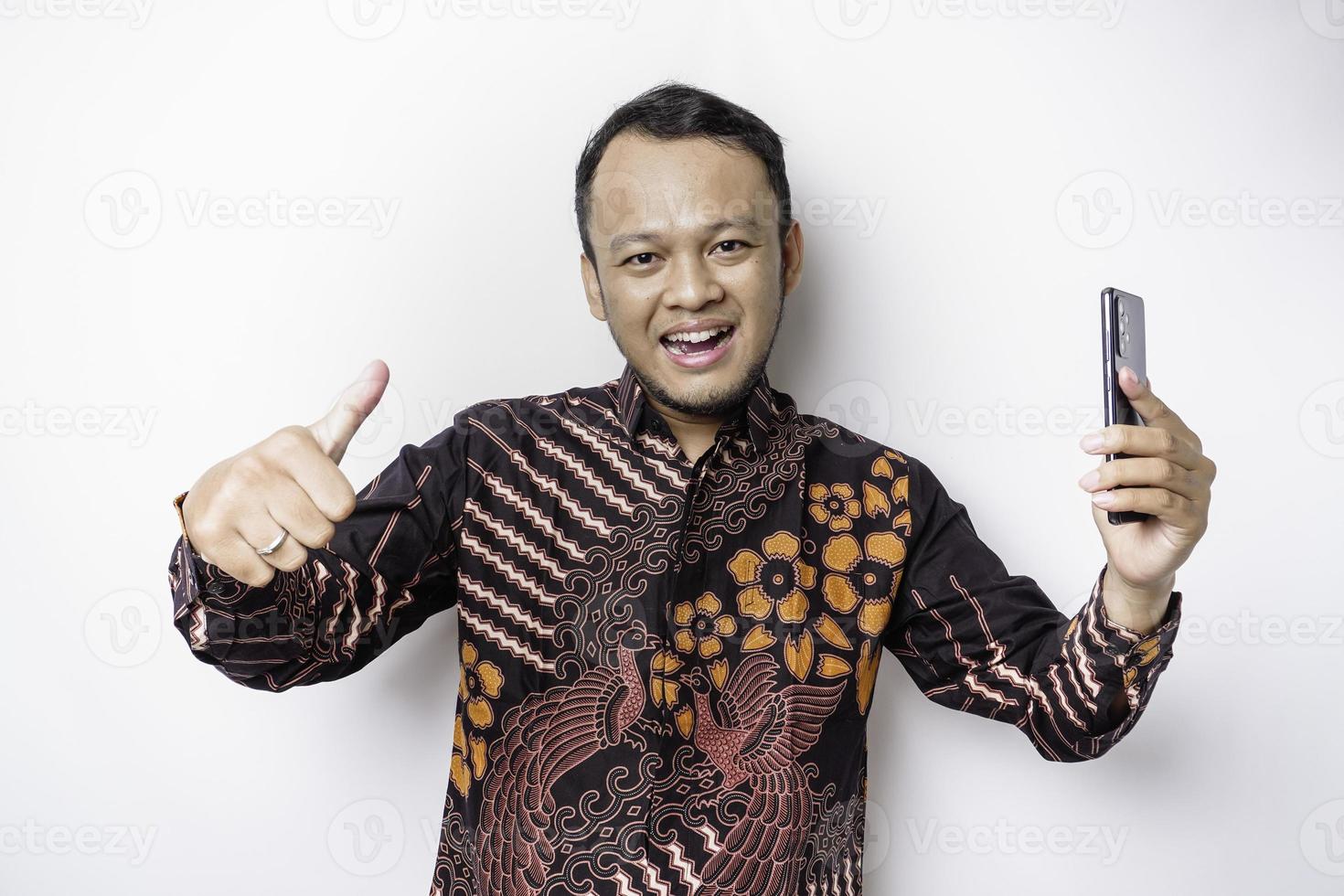 A portrait of a happy Asian man wearing batik shirt and holding his phone, isolated by white background photo