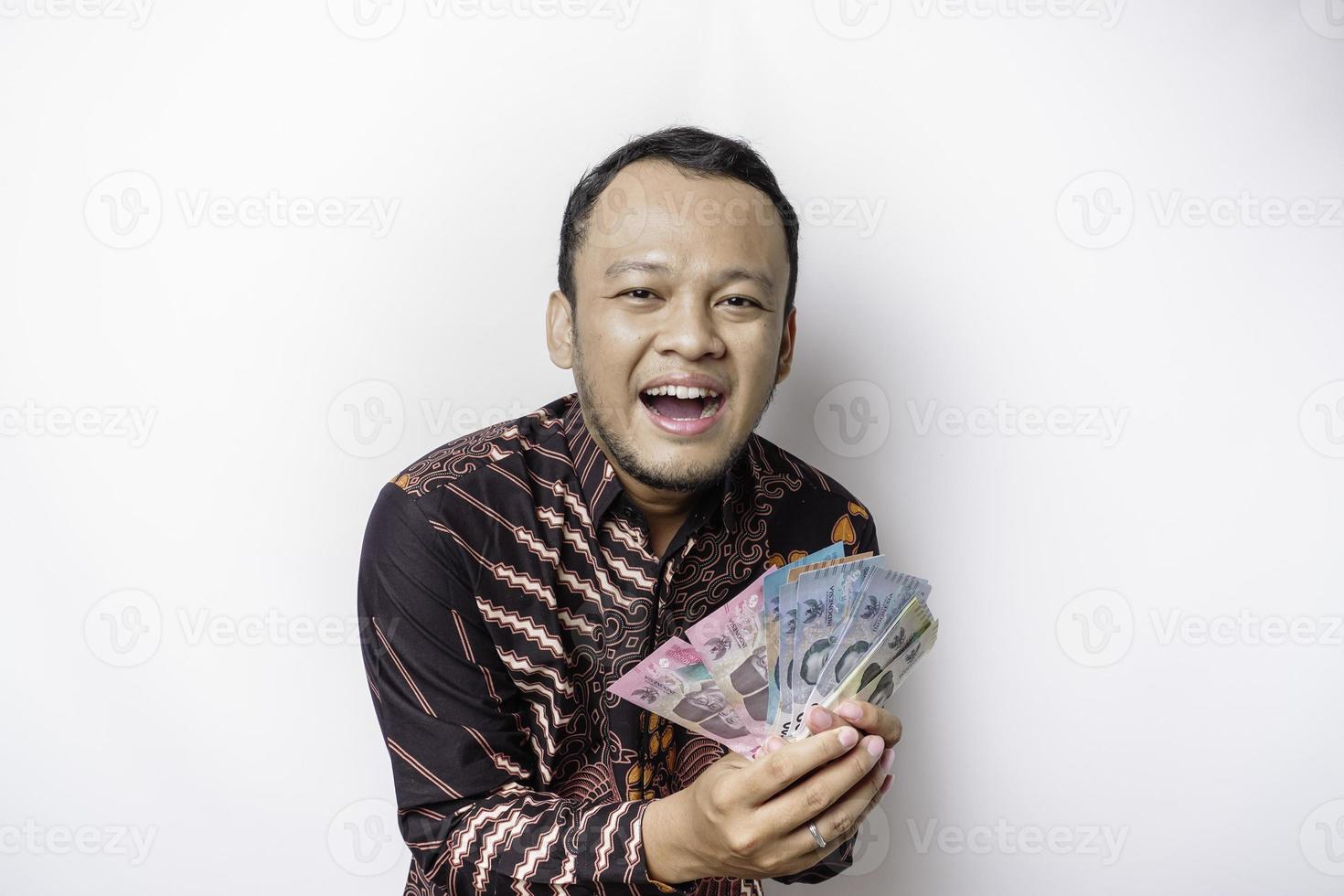 A happy young Asian man is wearing batik shirt and holding cash money in Indonesian rupiah isolated by white background photo