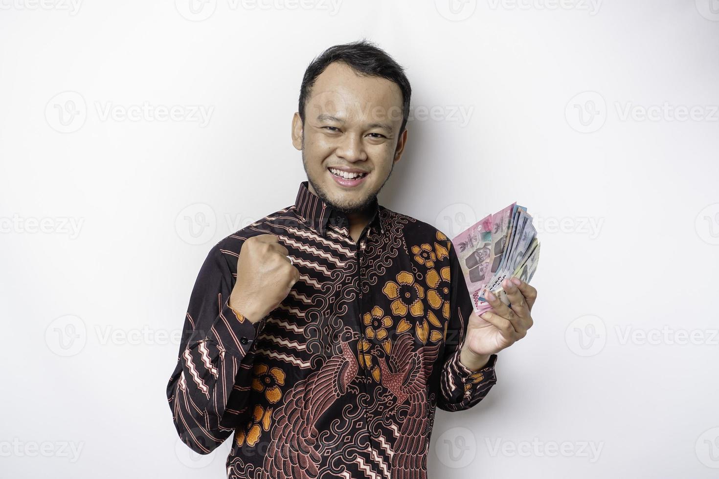 A happy young Asian man is wearing batik shirt and holding cash money in Indonesian rupiah isolated by white background photo