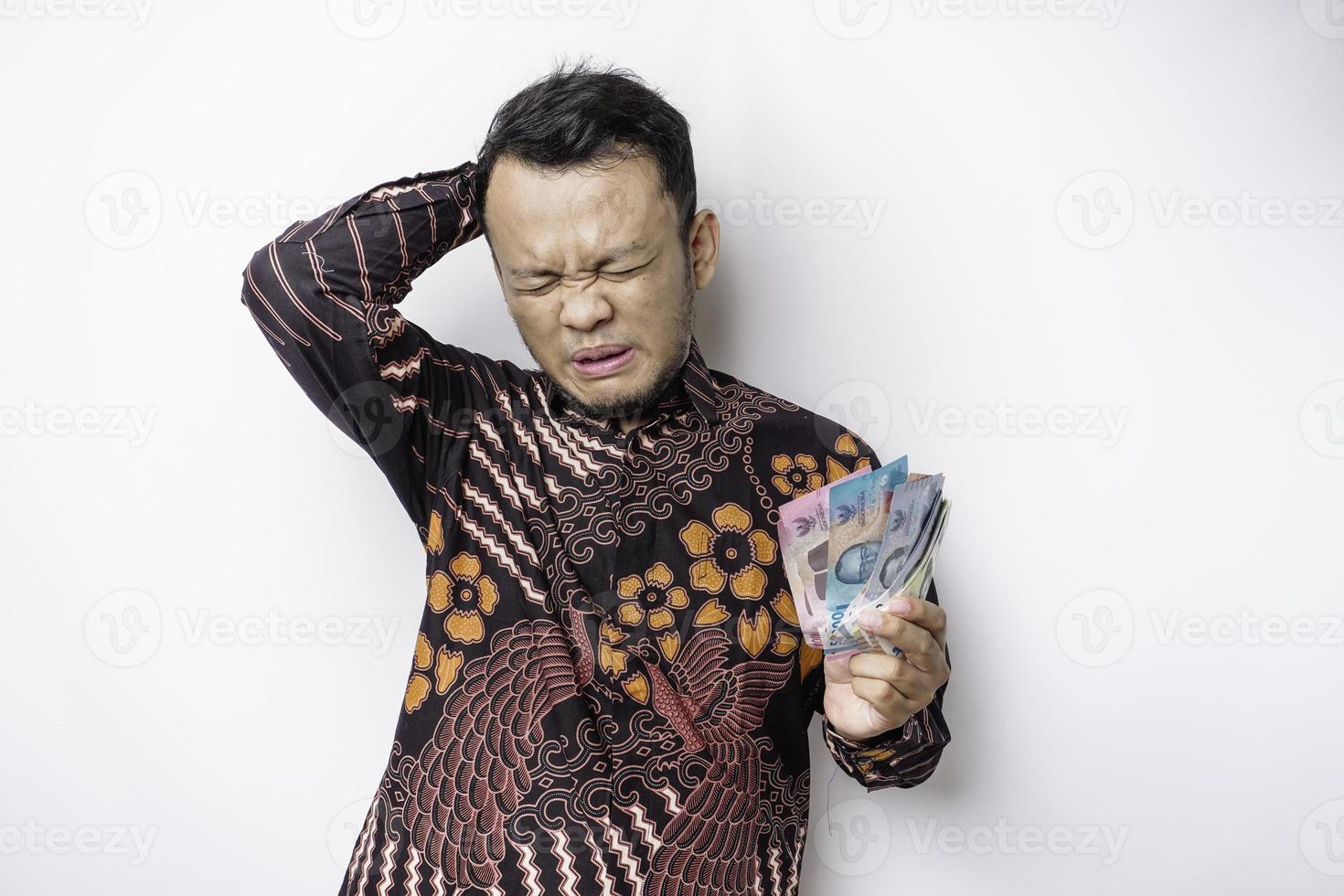 A stressed Asian man wearing batik shirt and holding money in Indonesian Rupiah isolated by white background looks depressed photo