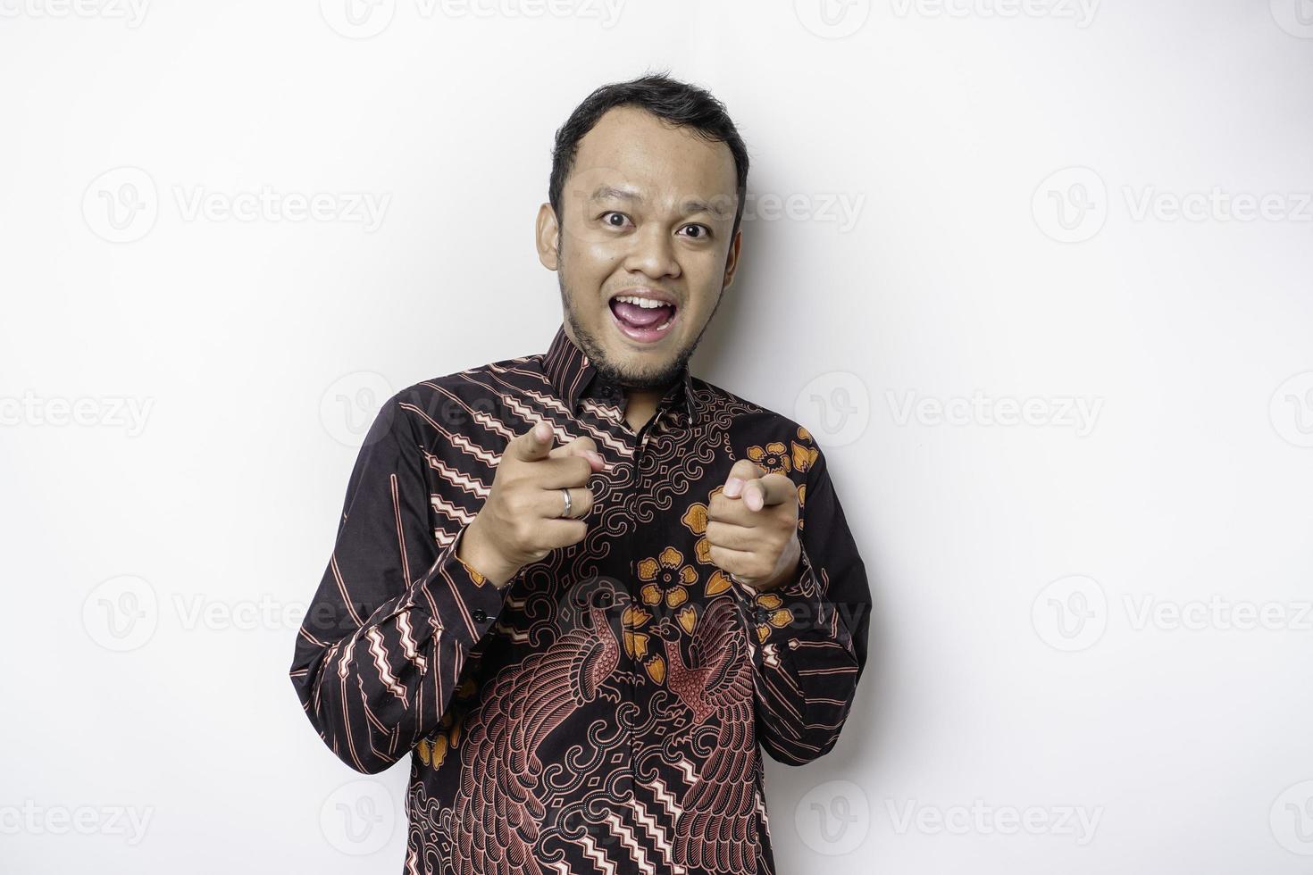 Young Asian man wearing batik shirt standing over isolated white background pointing fingers to camera with happy face. Good energy and vibes. photo