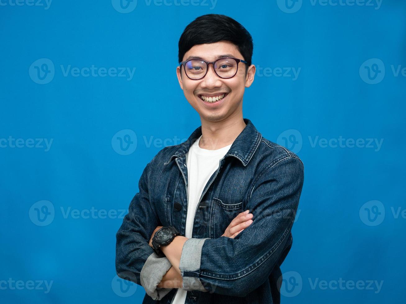 Young asian man jeans shirt cross arms gentle smile portrait blue background photo