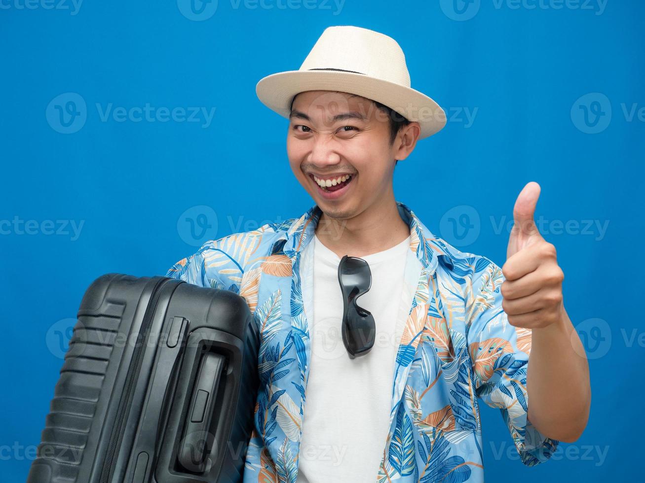 Positive traveler man cheerful hold luggage and thumb up blue background photo